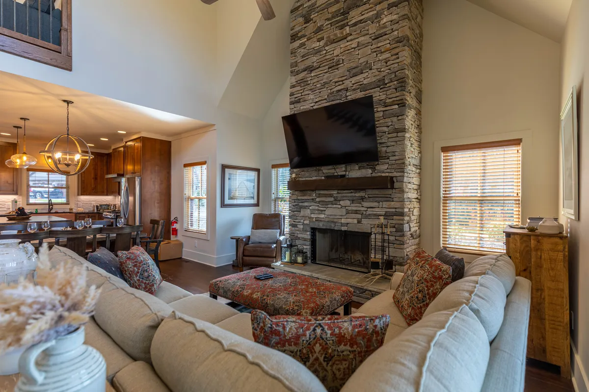 This image showcases a spacious interior with a grand stone fireplace as the central focal point, a large plush sectional sofa, a flat-screen TV mounted above the fireplace, and a wooden dining table situated nearby. The space is open-concept, allowing seamless flow between the living and dining areas, and the cozy atmosphere is enhanced by warm lighting and rustic-modern décor.