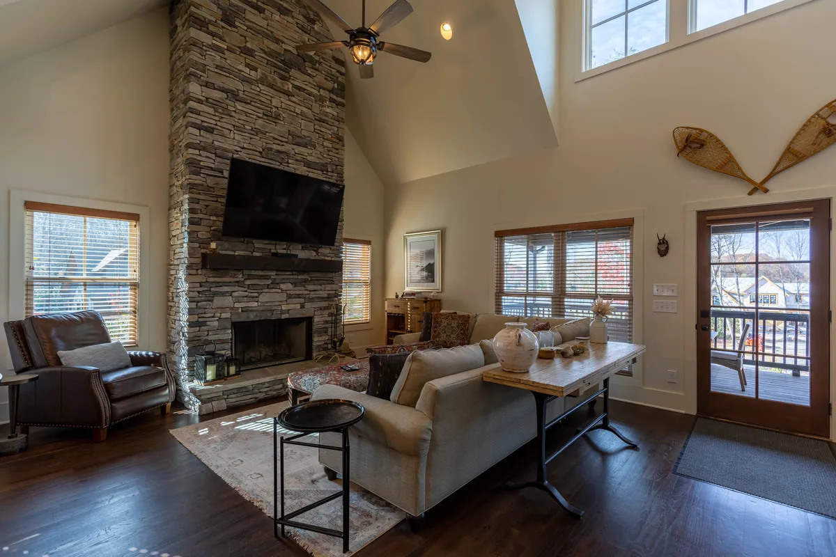 A spacious living area featuring a grand stone fireplace, expansive windows that flood the room with natural light, plush comfortable seating, and a large wooden dining table. The scene suggests a welcoming environment for group gatherings and shared meals.