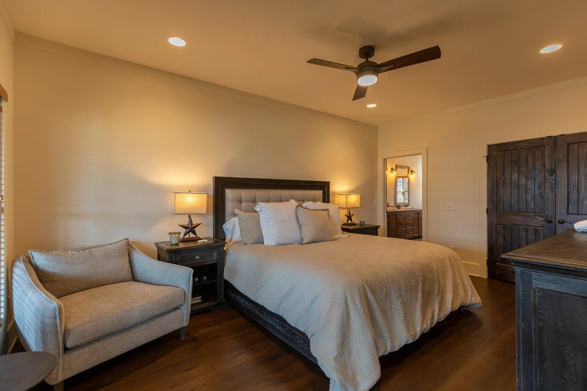 This image showcases a tranquil bedroom with a comfortable bed, nightstands, an armchair, and a wooden door. Natural light adds to the peaceful ambiance, and the decor exudes warmth and relaxation.