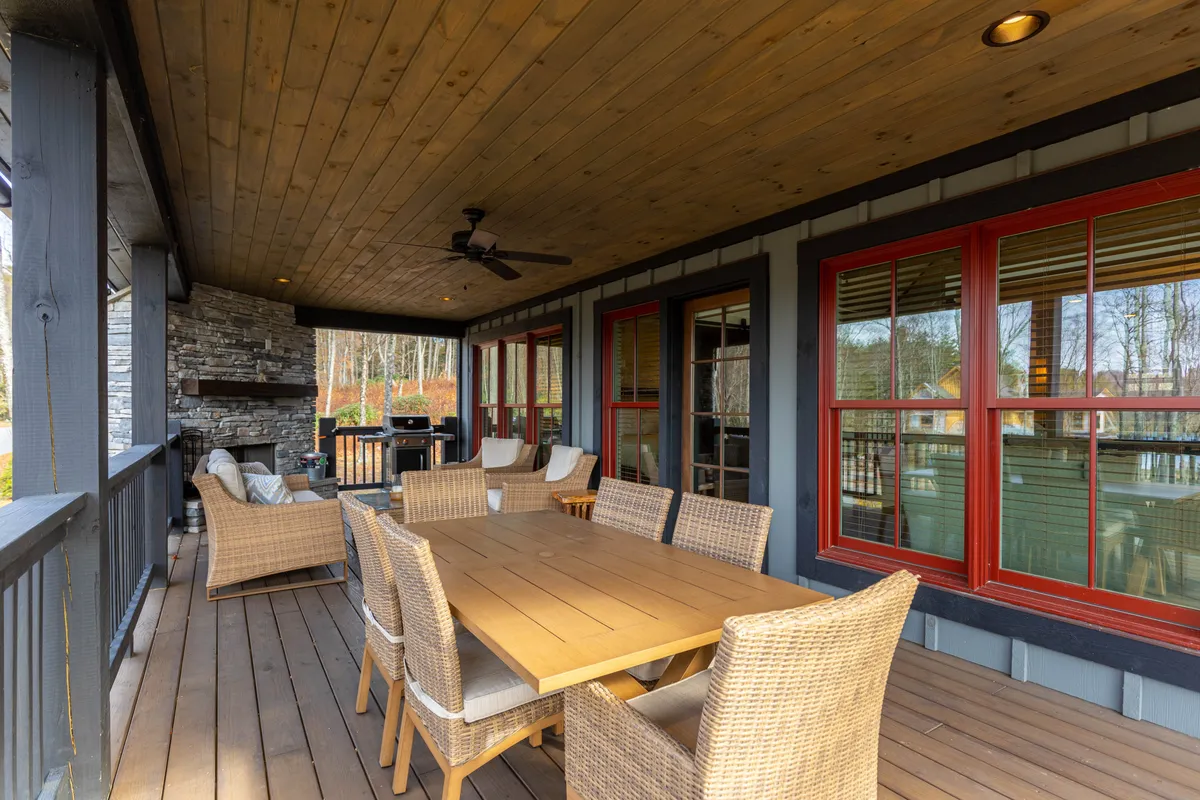 This image features a spacious wooden dining table surrounded by woven chairs on a back deck. A substantial stone fireplace serves as a focal point, complemented by large windows that hint at the adjoining indoor space. The deck appears inviting for both dining and lounging, with ample seating and a seamless transition from the indoors to the outdoors.