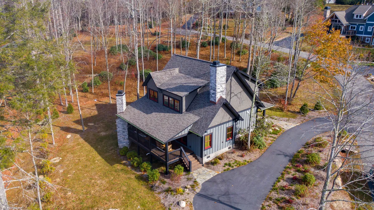 This exterior shot displays the home's striking combination of stone and wood siding nestled among mature trees. The driveway leads up to the house, and the porch glows softly in the sunset. The inviting outdoor spaces suggest peaceful morning coffees and lively evenings by the fire, with hints of the porch’s central role in mountain living.