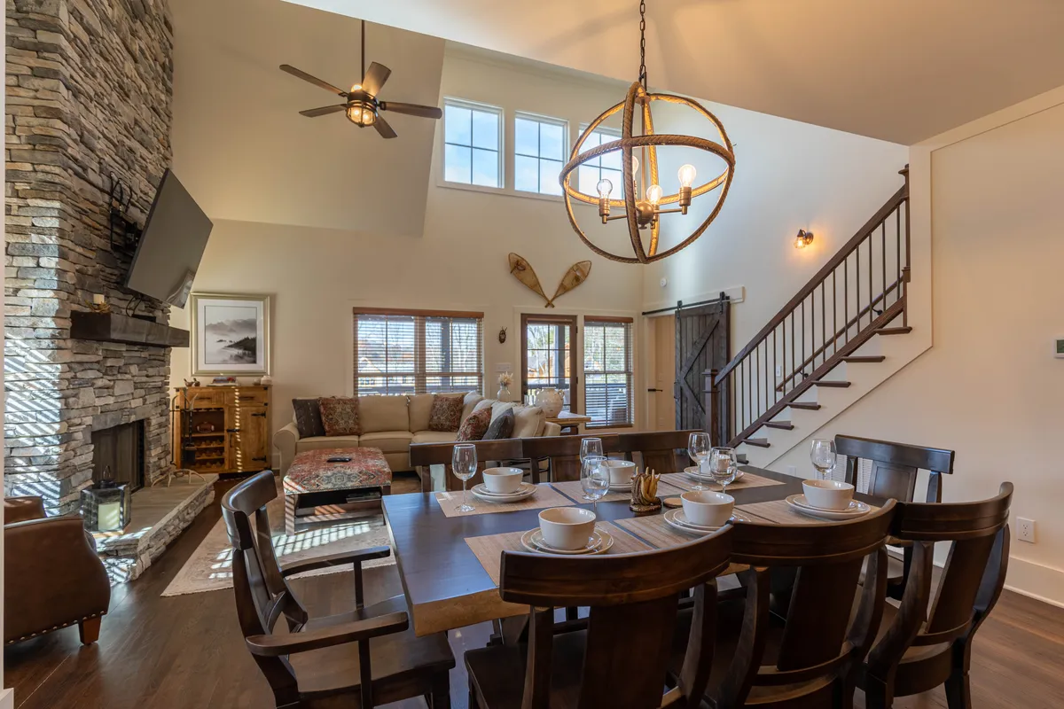 This image captures a spacious and warm living area with a dining table set for a meal, a comfortable sofa adorned with decorative pillows, and a striking stone fireplace as the focal point. The visible openness and natural light suggest a seamless connection to the outdoors, inviting guests to relax, gather, and enjoy the ambiance after a day spent exploring the mountains.