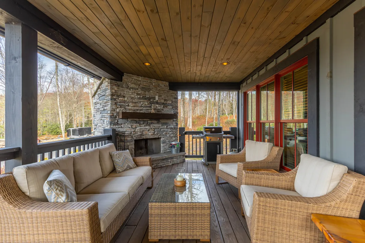 A serene sitting area centered around a rustic stone fireplace, complemented by woven furniture and a glass coffee table. Large windows offer views of the outdoors, blurring the line between the interior and the natural surroundings.