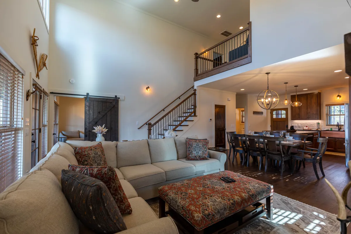 This image provides another perspective on the open living and dining area, showing a comfortable sectional sofa with decorative pillows, a dining table with chairs, and a staircase leading to the upper floor. The setup invites relaxation, conversation, and togetherness, while the arrangement demonstrates the seamless integration of common spaces.