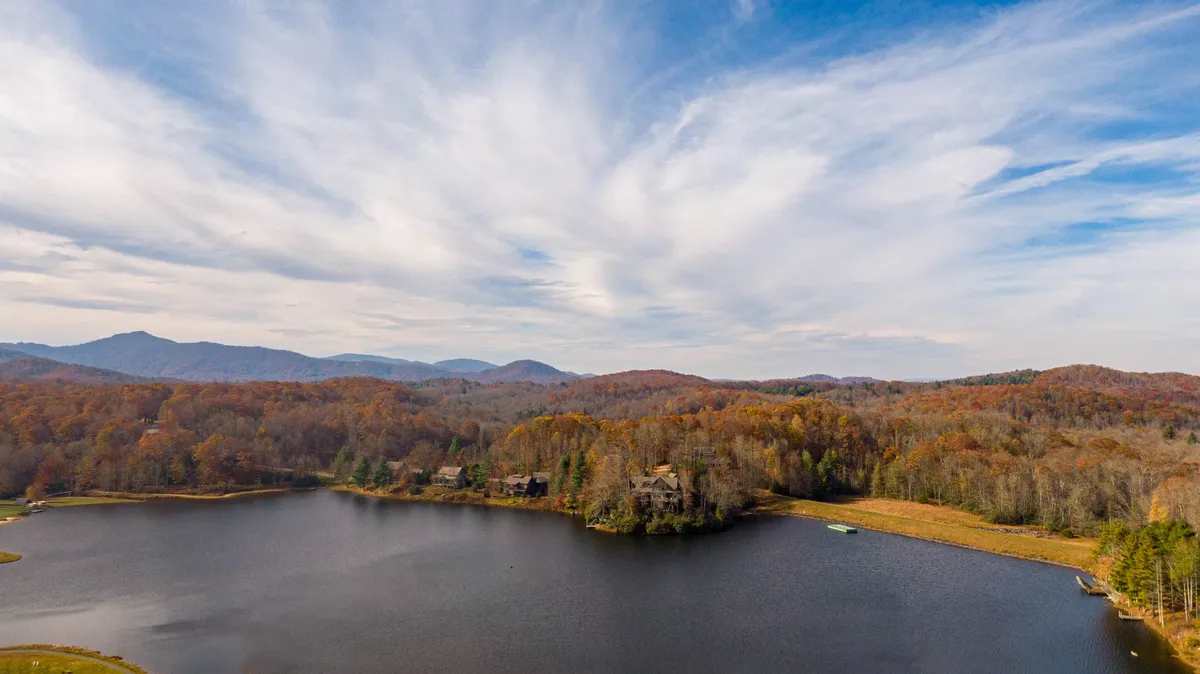 A serene lake surrounded by vibrant autumn foliage and distant mountains, with small cabins nestled in the scenery. The calm water invites activities like boating and fishing, while nature trails wind through the trees, hinting at outdoor exploration opportunities.