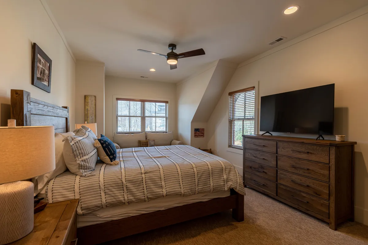 A cozy bedroom featuring a plush bed, dresser, television, windows letting in natural light, and a lamp. The room is inviting, with neutral tones and a relaxing vibe—ideal for both comfort and entertainment.