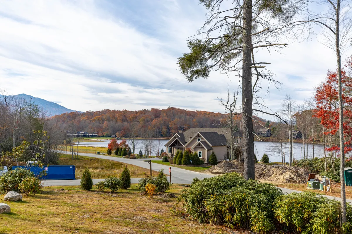 A serene exterior shot featuring a sparkling lake surrounded by lush trees, with houses nestled along the shoreline and mountains rising in the background. The scene highlights open water, gentle hills, and trails that wind through the landscape.